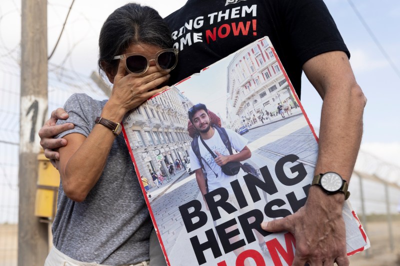 A woman at a rally wipes at her eyes behind a pair of sunglasses while holding up a poster at a rally. The poster reads "Bring Hersh home," Hersh being the name of her son.