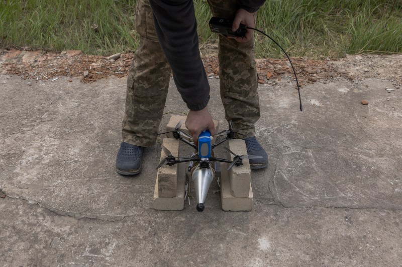 A small armed drone is seen on the ground between the feet of a soldier.