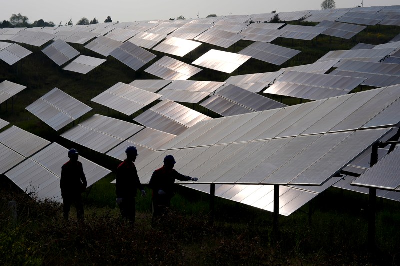 The silhouettes of three workers are seen from behind as they stand in front of a field of solar panels. One worker has a gloved hand extended to reach the nearest panel. Sunlight glints on the surface of panels farther on in the field.