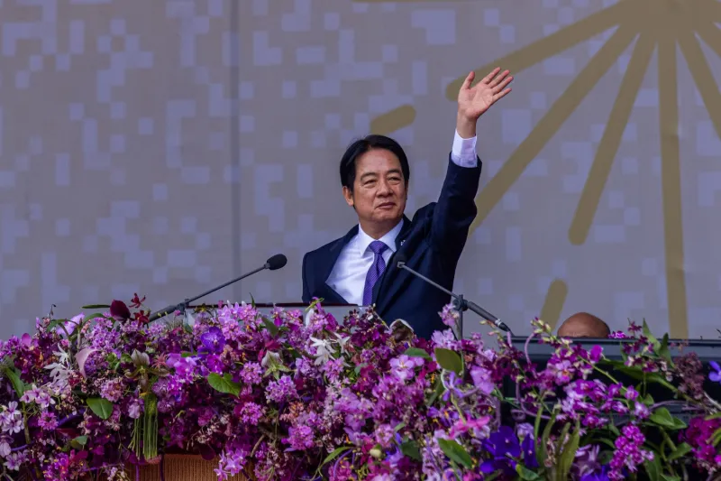 Taiwanese President Lai Ching-Te lifts one arm to wave while he stands in front of microphones at a podium decorated with an abundance of purple flowers. He wears a suit with a matching purple tie.