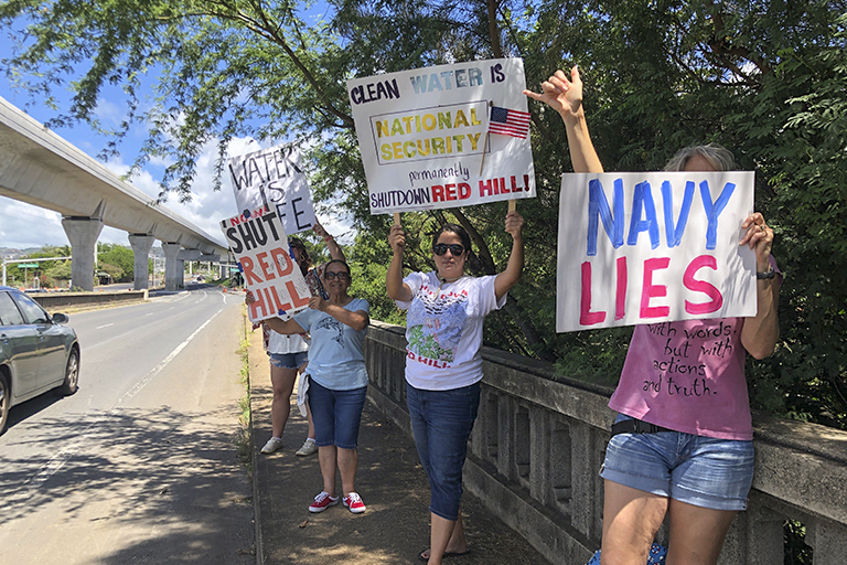 FILE &#8211; Protestors upset with the Department of Defense&#8217;s response to the leak of jet fuel into the water supply hold signs outside the gate at Joint Base Pearl Harbor-Hickam, Hawaii, Friday, Sept. 30, 2022. Three active-duty military members are taking the first step toward suing the U.S. government over jet fuel that contaminated drinking water in Hawaii. Their attorneys say the pre-litigation claim forms will allow them to later file a federal lawsuit in Honolulu. In 2021, jet fuel spilled from a drain line at a storage facility that flowed into a drinking water well and into the Navy&#8217;s water system. (AP Photo/Audrey McAvoy, File)