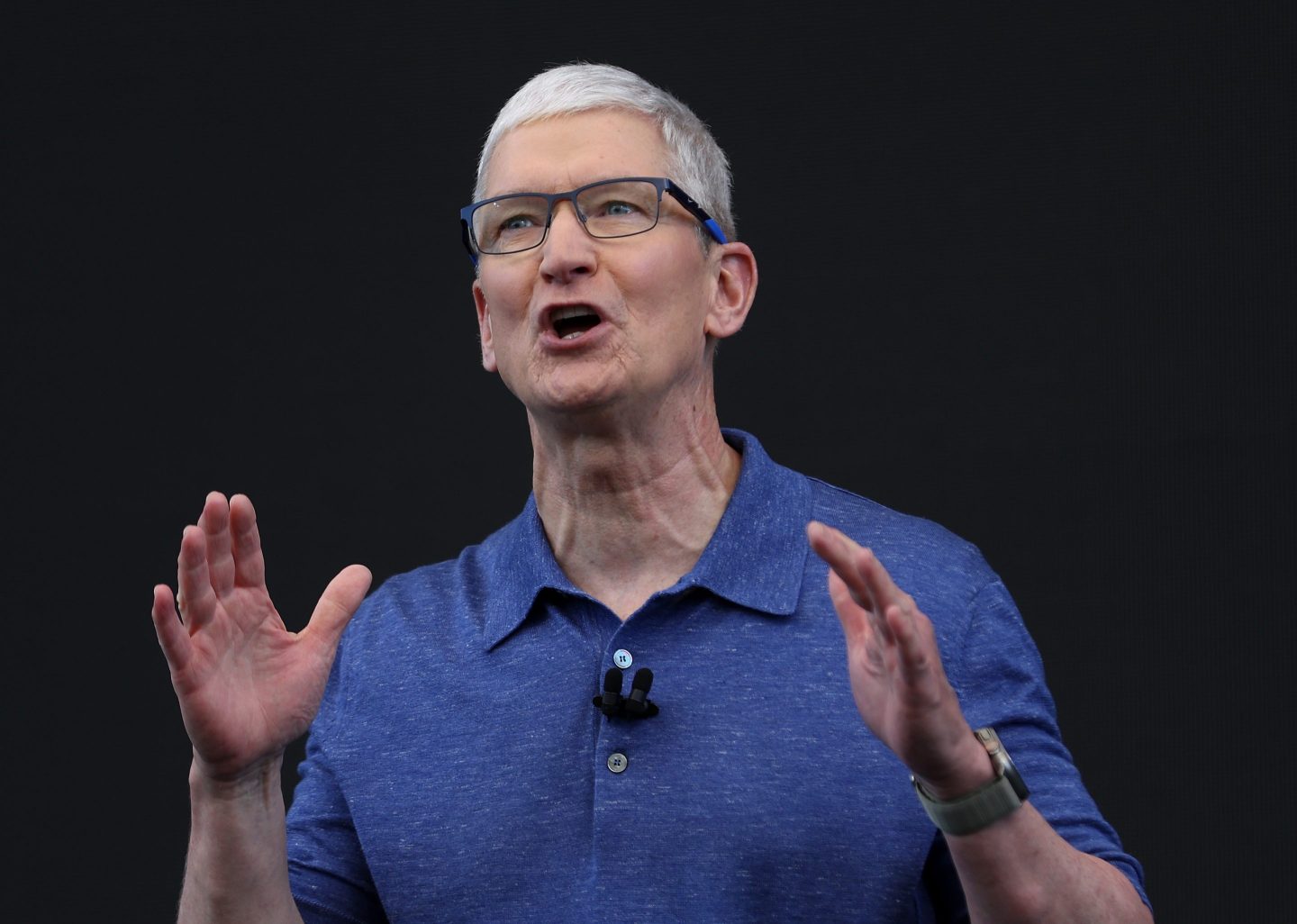 Apple CEO TIm Cook wearing a blue T-shirt and raising his hands while giving a speech.