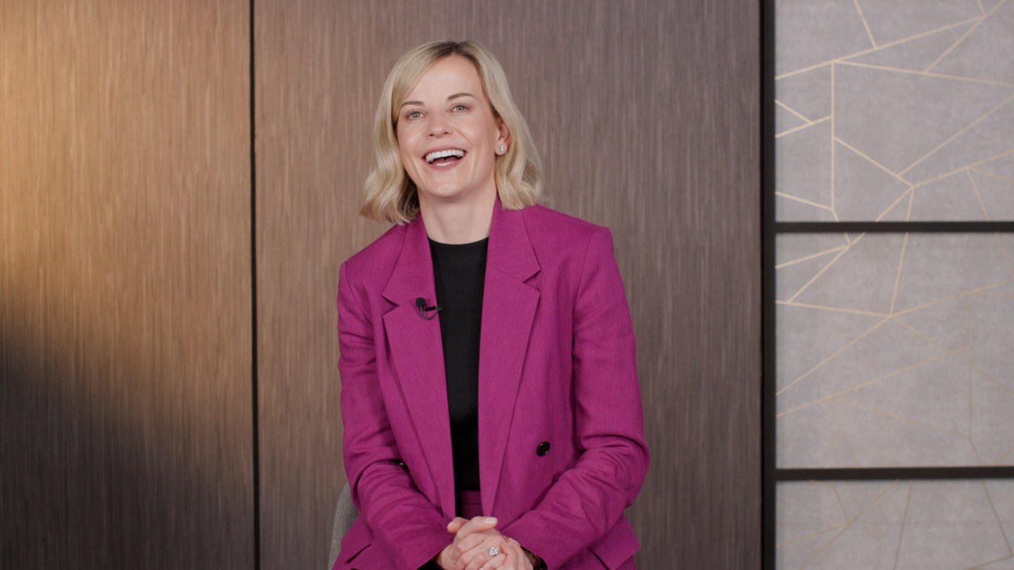 Susie Wolff, wearing a pink suit, smiles in front of a brown background.