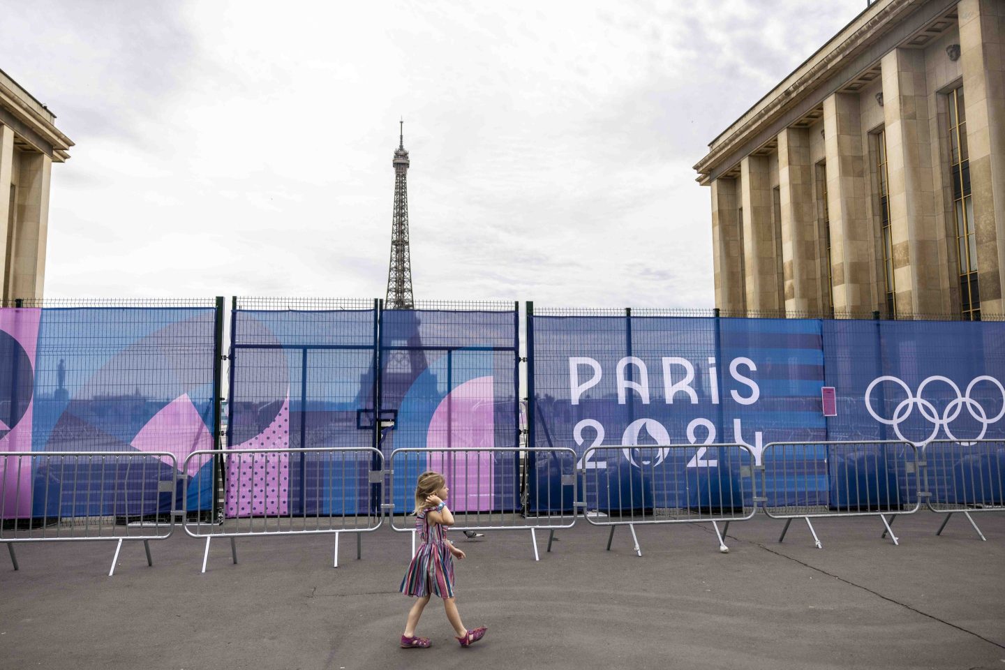 A girl walks past a baracade with the Eiffel Tower and a sign for the Paris Olympics in the background.