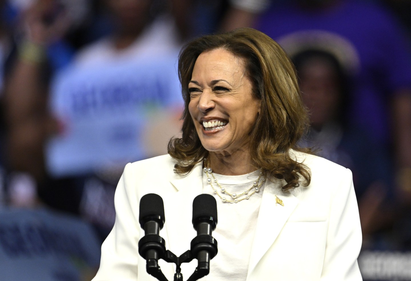 U.S. Vice President Kamala Harris, dressed in a white blazer, smiles broadly while speaking at a podium with two microphones