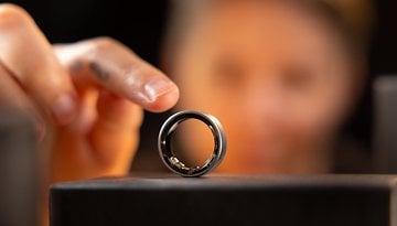 A close-up of a Samsung Galaxy Ring on a stand, with a hand reaching to touch it in the background.