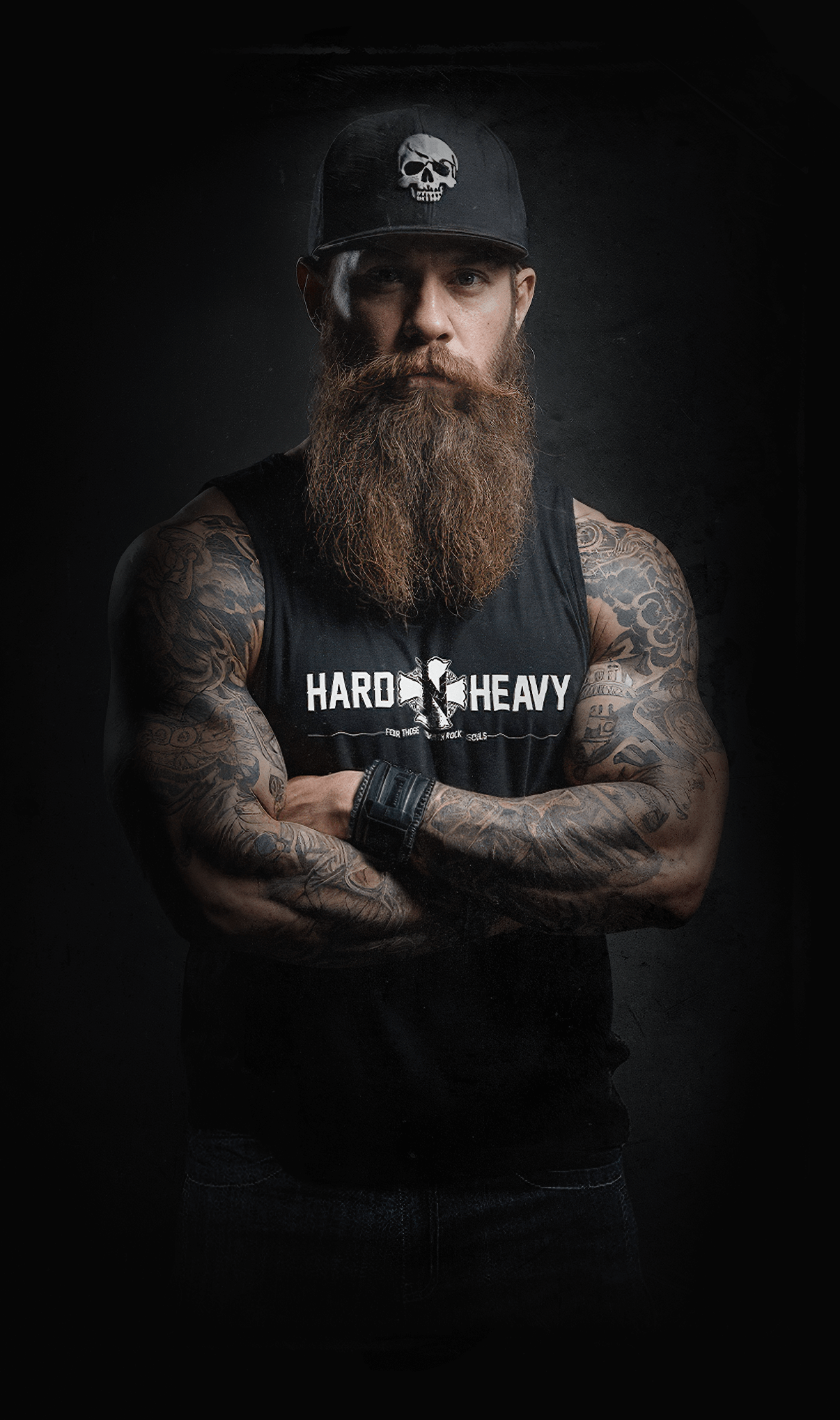 Bearded man in HARD'N'HEAVY tank top and cap, arms crossed, against a dark backdrop.