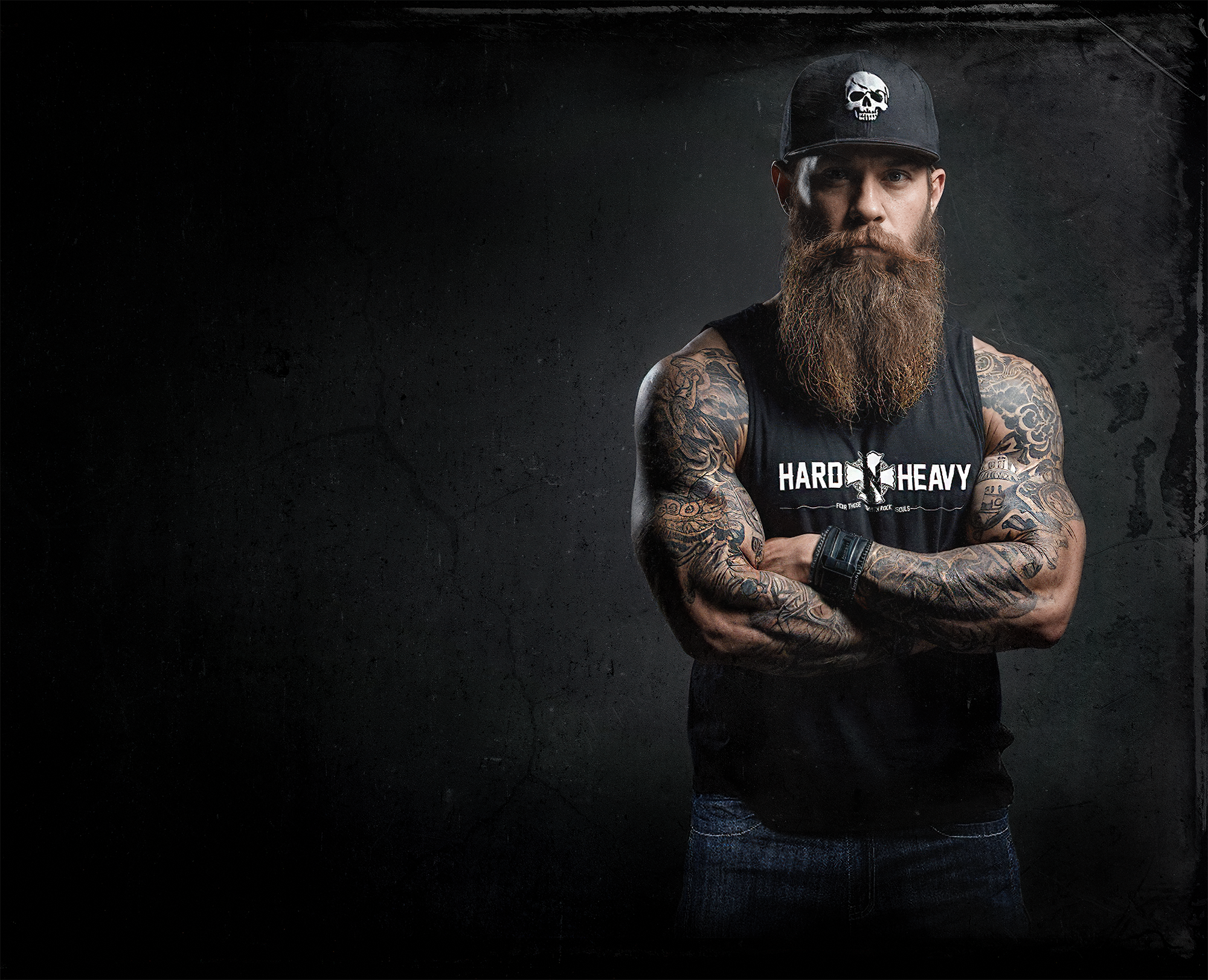 Bearded man in HARD'N'HEAVY tank top and cap, arms crossed, against a dark backdrop.