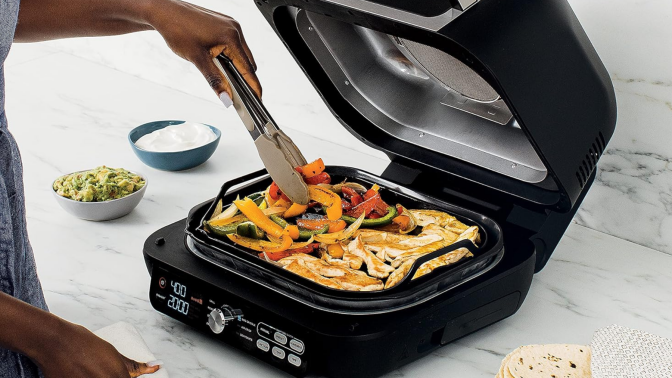 Person's hand using tongs to cook vegetables and chicken on Ninja griddle