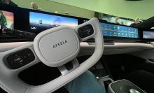 A close-up view of the interior of an AFEELA car, focusing on the futuristic steering wheel with "AFEELA" branding. The dashboard features a wide digital display with driving information and an app interface.