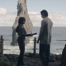 Osha holds an unlit lightsaber between her and the Stranger; the two stand at a cave entrance overlooking the ocean.