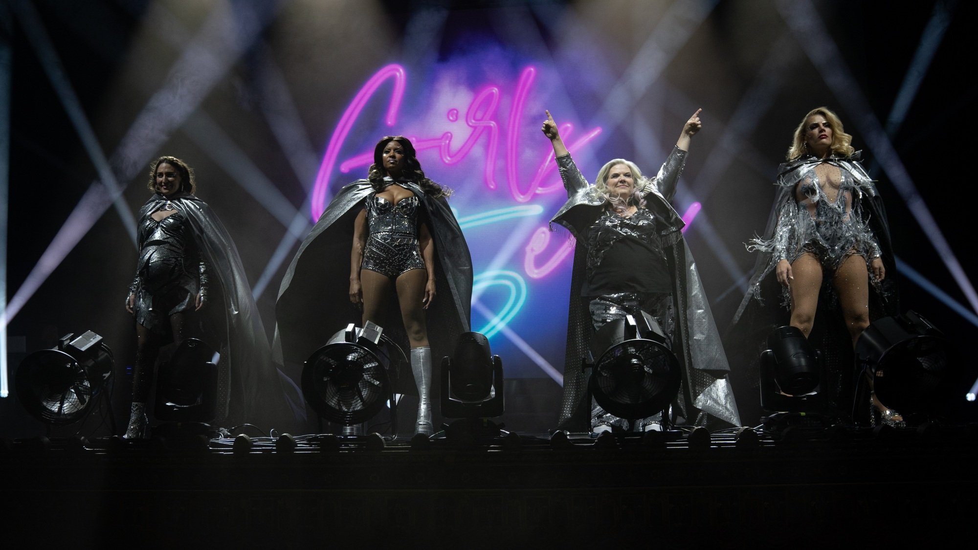 A band of four women stand onstage in elaborate silver outfits against a backdrop that says "Girls5eva."