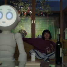 A large white robot with a glowing face and a human woman sit on a couch watching TV.