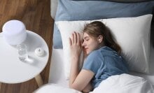 an overhead view of a person sleep in bed while wearing a sleep earbud which a table beside her has the earbud case and a glass of water