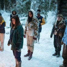 A group of girls stand in the snow outside a cabin in the woods.