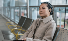 a person with french braided hair sits on a chair in a row of chairs while holding a phone in the left hand and using a sony WF-1000XM5 earbuds in the left ear