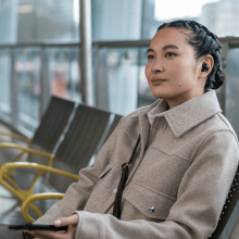 a person with french braided hair sits on a chair in a row of chairs while holding a phone in the left hand and using a sony WF-1000XM5 earbuds in the left ear