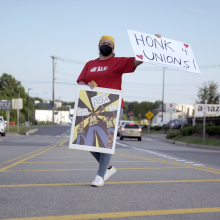 An ALU member stands in the median of a road holding a sign that reads "Honk 4 Unions."