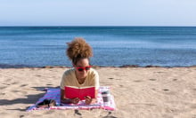 a person lays on a beach towel on the beach reading a red book with the ocean behind her