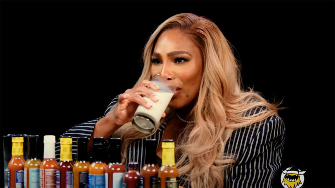 A woman sits at a table covered in sauce bottles, drinking a glass of milk.