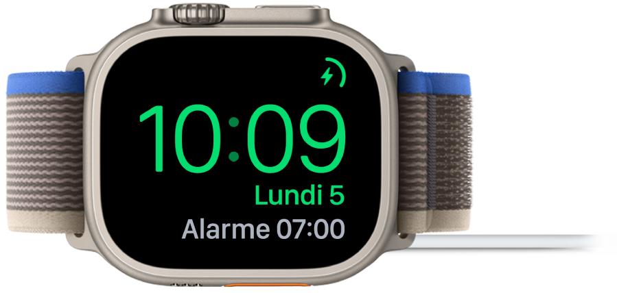 Apple Watch placée sur sa tranche et connectée au chargeur. L’écran montre le symbole de recharge dans le coin supérieur droit, l’heure actuelle en dessous et l’heure du prochain réveil.