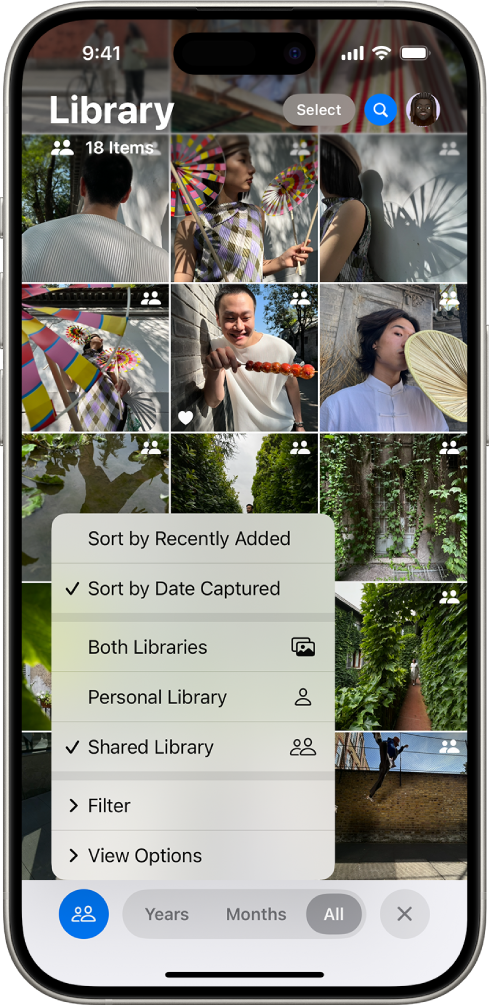 An iCloud Shared Photo Library in the Photos app. The Shared Library button in the bottom-left corner is selected, and Shared Library is selected in a list of library options. All the photos displayed in the library have a shared library badge in the top-right corner.