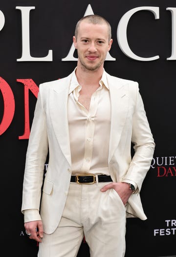 joseph quinn smiles for the camera wearing a cream open neck shirt and suit