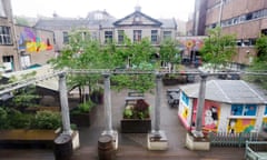 An urban square in front of a grey stone Victorian institutional building, given extra colour by a mix of leafy trees and mural art on different walls and buildings