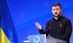 Volodymyr Zelenskiy speaks at the conference: he stands against a bright blue background with the blue and yellow of the Ukrainian flag seen in one corner, and wears a black jumper; he has one hand raised and looks solemn.