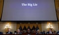 five people sit at a desk beneath a big screen with the words 'the big lie'