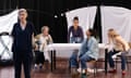 Five women on stage. One stands at the front of the stage looking out while the other four stand and sit around a table with a white tablecloth