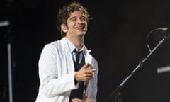 Man wearing white suit jacket and black tie smiles and holds a microphone on stage