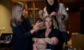 Hannah Neeleman holds a baby as two other women do her hair and makeup