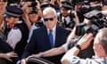 Huw Edwards wearing sunglasses and a suit walks towards a car with an open door surrounded by photographers as police try to clear a way for him