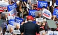 A man in a black suit and red baseball cap is seen from behind, with a sea of people cheering and holding red, white and blue Trump 2024 signs taking up most of the frame.