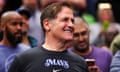 a man wearing a black T-shirt reading 'Mavs' smiles while watching a basketball game