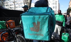 The back of a Deliveroo rider, surrounded by riders from Just Eat and Uber Eats, on a bicycle wearing a blue Deliveroo backpack.