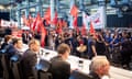 A row of executives in dark suits sit at a long table in the foreground while, in front of them, factory workers hold banners