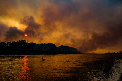 Black smoke darkens a red sky above a body of water against a black tree line