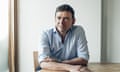 The author David Nicholls sits at a wooden table in a blue button down shirt.