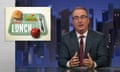 John Oliver, wearing a dark suit and glasses, sits at a desk and speaks with a graphic of of a school lunch tray on the screen