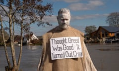 Man in hessian smock with white-painted face stands in front of water with sign around his neck reading: 'Thought greed was good - it turned out crap'