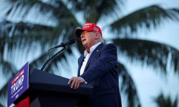 white man wearing blue suit and red hat stands at podium in front of microphone with palm tree in the background