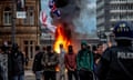 People, some in hoods and balaclavas, stand in front of flames with St George's flag and Union Jack in background and police officer in foreground