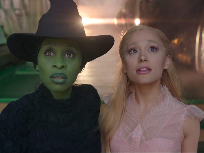 cynthia erivo and ariana grande as elphaba and glinda in wicked. erivo is painted green and wearing a black dress and hat, while grande has blonde hair and is wearing a pink dress