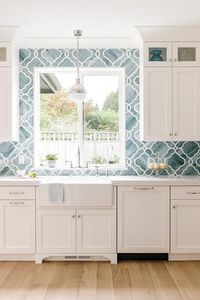 Glass front display cabinets are stacked over white cabinets mounted on either side of a window framed by blue quatrefoil backsplash tiles.