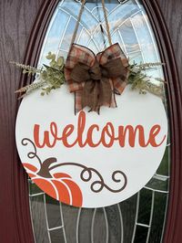 Add a touch of fall to your front door with this "Welcome" round wooden door hanger featuring an adorable pumpkin!  Comes in your choice of two sizes, 15" or 18" round.  Complete and the beautiful bow,  greenery and attached rope for hanging. Wood round details: -Choose your size- 15 inches round (as shown is pictures) or 18 inches round -Greenery and bow are included and attached with special adhesive and staples -Rope is attached on the back for easy hanging -Wood is sealed for protection but