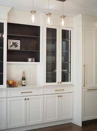 Three clear glass mini pendants hang in front of glass front white cabinets contrasted with dark stained wood interiors and positioned flanking dark stained wood shelves.