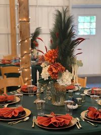 Set up beautiful tablescapes at your event using an attractive orange, green, & gold color palette. Drape the tables with emerald green tablecloths & flaunt orange & white hydrangeas & white amaranthus sprays, along with green plumes in vases. Also, set gold mercury candle holders & use gold chargers, black plates, gold cutlery, orange napkins, & gold rim clear flutes to glam up the place setting. Moreover, cover the venue pillars with string lights to enhance the ambiance.