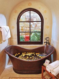 Tucked below an arched window, a freestanding copper tub transforms this bathroom into an elegant, relaxing retreat. The neutral tilework surrounding the tub and the delicate seashell border framing the window enhance the space's soothing atmosphere.